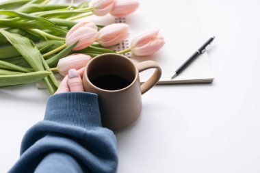 Masasında bir fincan kahveyle bir buket lale çiçeği ve bir not defteri olan bir kadın. Öğretmenler Günü. Açık pembe çiçekli laleler çiçek buketi ve beyaz masa arkasındaki boş defterleri aç.