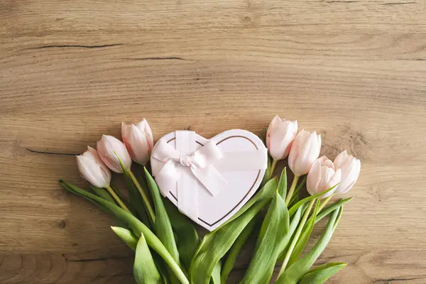 Taze pembe laleler ve kalp şeklinde bir hediye kutusu, tahta bir masanın üzerinde, düz yatıyordu. Sevgililer Günü, Kadınlar Günü ve Anneler Günü için fotokopi alanı olan tebrik kartı. Üst görünüm.