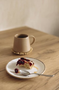 Sabahleyin mutfak masasında bir fincan kahve ve vişneli turta artığı. Sıcak ve sıcak bir atmosfer, kırsal ahşap dokular, huzurlu bir kahvaltı çağrıştırıyor ya da evde yavaş bir sabah anı..