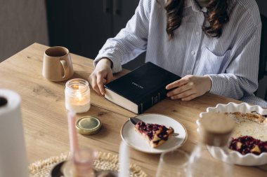 Sıcak bir mutfakta kahvaltı ederken İncil okuyan bir kadın. Ilık sabah ışığı, inancın, yansımanın ve günlük bağlılığın huzurlu bir atmosferini yaratıyor..
