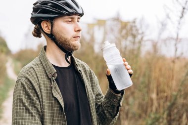 Günlük giysiler içinde bisiklet kaskı takmış su içen genç sakallı bir adam. Hidrasyon kavramı, aktif yaşam tarzı, açık hava fitness ve sağlık.