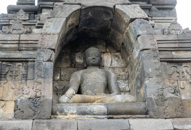 Bir Buda heykeli, Borobudur Tapınağı 'nda antik oymalarla çevrili bir taş oyukta sakince oturur..