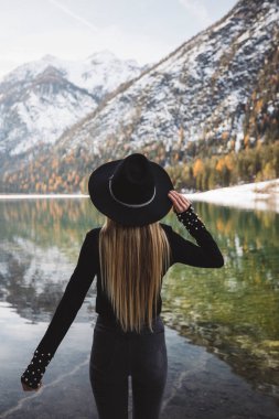 Back view of woman in black hat standing on the lake shore and watching the mountains  