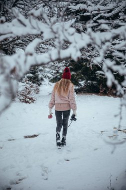 Back view of blonde woman with camera and red cup in hands walking on the snow covered forest 