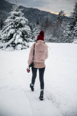 Back view of blonde woman with camera and red cup in hands walking on the snow covered forest 