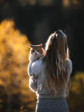 Back view of young blonde female holding a cat in her arms. People and domestic animal relationship concept. Autumn season 