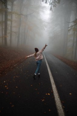 Young female with raised hands walking on the road in the foggy forest, back view 