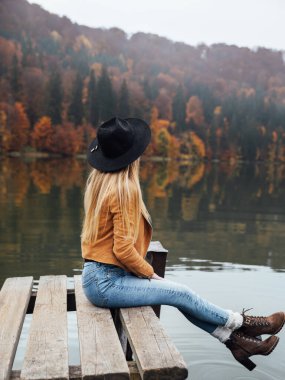 Side view of blonde woman wearing brown jacket and hat sitting on a wooden pier near the volcanic lake in autumn season 