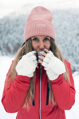 Young woman wearing winter clothing is enjoying hot tea from mug outdoors. Travel concept. Keeping warm in cold weather