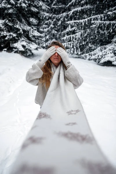 Magnificent girl in winter outfit posing in winter forest. Stylish european woman with long knitted scarf hiding her face with the hands.