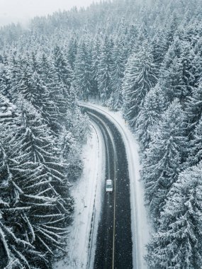 White car drives empty road in forest in the cold winter. Tourists on road trip cruising through the idyllic snow covered countryside and woods. clipart