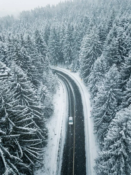 White car drives empty road in forest in the cold winter. Tourists on road trip cruising through the idyllic snow covered countryside and woods.