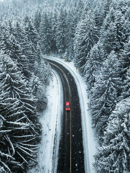 Tourists on road trip cruising through the idyllic snow covered countryside and woods. Red car drives empty road in forest in the cold winter.