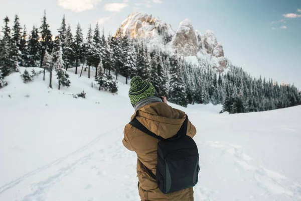 Male photographer in warm clothes taking photos with the camera from a snow covered trees and mountains at sunrise.