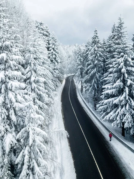 Snowy winter road in the mountains. Aerial view of a beautiful snow covered  forest and a hiker woman on the road. Travel concept.