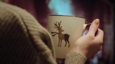 Over the shoulder young woman relaxing near fireplace with a white cup of tee in cozy christmas woolen sweater. High quality 4k footage