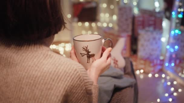 肩越しの若い女性が居心地の良いクリスマスにお茶を飲みながらリラックスしていると バックグラウンドショットで装飾されたリビングルームのある靴下とセーターが盗まれました 高品質4K映像 — ストック動画