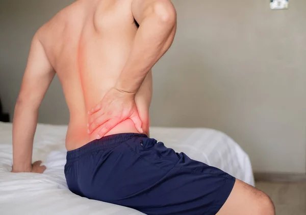 The man sat on the white bed, holding his aching waist as he was about to get up. Inflammation of the lumbar region. Twisting. Getting up in the wrong position. Inflammation of the muscles of the back