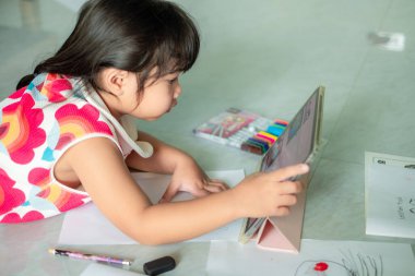 Asyalı kız, 5-6 yaşlarında, video kliplerle origami yapıyor ya da evde tablet, uzaklık öğrenimi, evde eğitim, iş formu ile internet üzerinden çalışıyor.