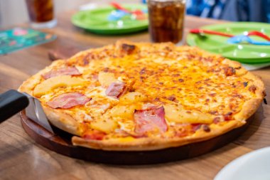 Eritilmiş peynirli, jambonlu ve ananaslı taze fırınlanmış Hawaii pizzası, arka planda içecekler ve renkli sofra takımlarıyla ahşap bir tabakta servis ediliyor.