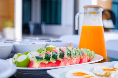 Dilimlenmiş karpuz, guava ve salatalıkla birlikte beyaz bir tabakta sunulan renkli kahvaltı seti, yanında bir sürahi taze portakal suyu ve güneşli yan yumurtalar.