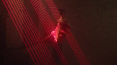 Female dancer in short stage pink dress of Latin American ballroom dancing is circled in dance support on outstretched arms of man. Several parallel beams of red laser light in dark. Performance part