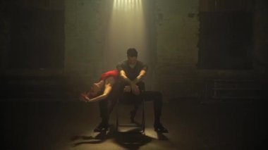 Empty loft room,young man sits on chair in center,woman starts dancing around,waking him from sleep.Latin-American ballroom dance.Gradually additional light beams turning on.Cinema art atmosphere