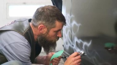 Sakallı, beyaz saçlı, orta yaşlı bir usta silikon yuvarlak numune kullanarak düzgün kıvrımlı araba gövdesini fırçalıyor. Otomobilin siyah arka planında dalgalı beyaz hava akışları çiziyor