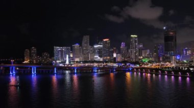 Gece yavaş çekim panoramik manzara Miami City, Florida 'nın deniz manzarası, şehir merkezinin ışıkları gökdelenler aydınlandı karanlık bulutlu gökyüzü, renkli yansımalar ve sakin sularda yüzen tekne.