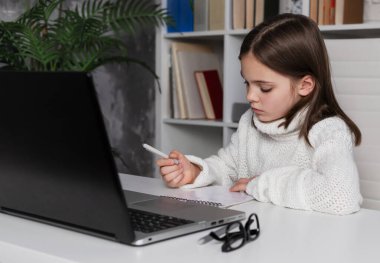 Ciddi esmer okul çocuğu evde ders çalışıyor. Kız dizüstü bilgisayarla masada oturuyor, deftere yazıyor, ödev yapıyor ya da sınava hazırlanıyor, online eğitim kavramı, e