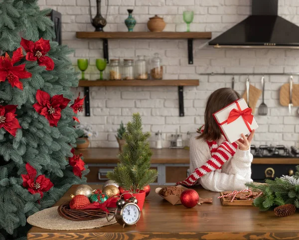 Kız elinde kırmızı kurdeleyle beyaz hediye kutusunun arkasında saklanıyor. Çocuk Noel süslemeli mutfakta, ahşap masada oturuyor.