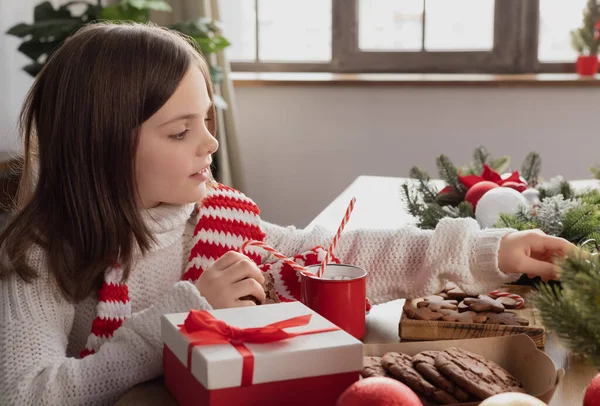 Kız ahşap masada oturuyor ve kırmızı kurdeleli, zencefilli kurabiyeli, Noel süslemeli kırmızı bardaklı beyaz hediye kutusuyla ilgileniyor.