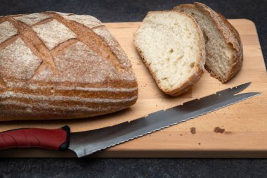 Oval somun taze pişmiş ekşi ekmek, tahta kesme tahtasına saplanmış bıçaklı ekmek, açı manzaralı. Koyu gri masada kırsal ekşi ekmek