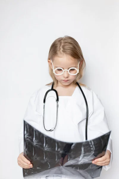 Caucasian child dressed up in doctor uniform with eyeglasses and stethoscope, looking thoughtful and serious. Kid holding and looking at x-ray film of radiographic, ct scan, MRI of spine