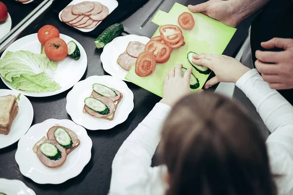 Top view, hands of father and daughter cooking breakfast. Sandwitches with vegetables, bacon and bread on dark table