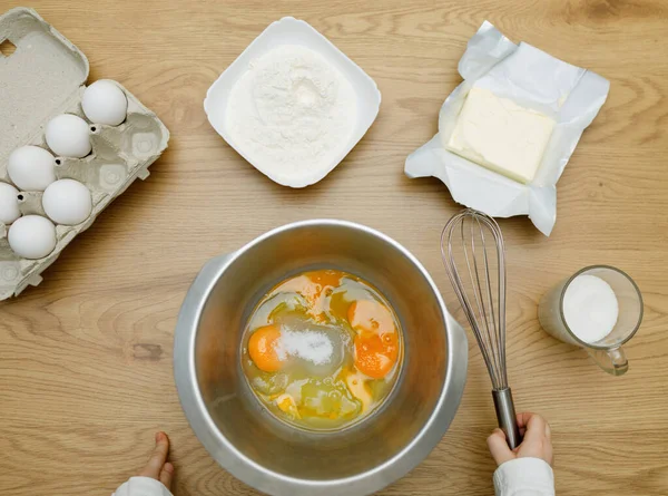 Kid's hands with mixing whisk. Eggs, sugar, butter, flour. Cooking ingredients in bowl, ready for mixing dough. Cookbook.