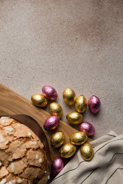 Easter dove cake or bread kulich with chocolate Easter eggs, wooden cutting board, striped napkin on concrete background. Vertical, place for recipe, copy space, top view