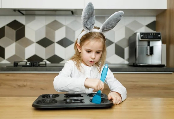 Tavşan kulaklı ciddi küçük kız modern ahşap mutfakta zeytinyağlı mavi silikon fırçayla pişirme kabını yağlıyor. Paskalya konsepti, ev yapımı yemekler, ebeveynlere yardım.