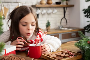 Bir fincan sıcak kakao ya da çikolataya üfleyen çocuğun yakın çekimi. Xmas 'ın dekore edilmiş mutfağında küçük bir kız. Beyaz örgü süveter ve çizgili eşarp giyen bir çocuk. Noel ve Yeni Yıl tatili