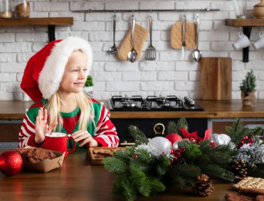 Elf gibi giyinmiş, şeker kamışı tutan ve gülümseyen bir çocuk. Zencefilli kurabiye, tepside çikolatalı kurabiye, tezgahta çelenk.