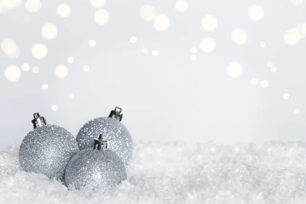 Three silver glitter Christmas ornaments with silver cap on top on bed of snow on blurred white background with bokeh effect of lights. Copy space
