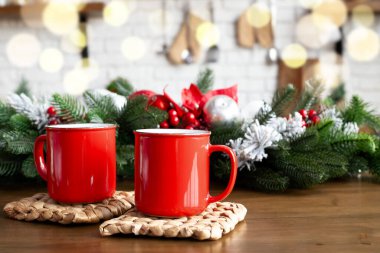 Ahşap masada iki kırmızı kupa ve mutfak arka planında bokeh ışıkları olan Noel süslemeleri. Tezgahta kırmızı meyveler ve mücevherlerle süslenmiş çam dalları.