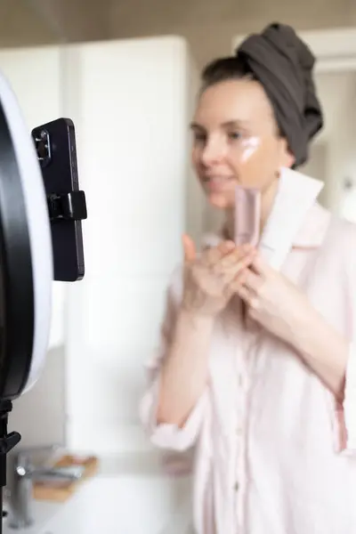 Güzellik ürünleriyle dolu şişeleri banyoda elinde tutan bir kadın akıllı telefona bakıyor. Günlük kaydı rutini, vloglama, kullanıcı tarafından oluşturulan içerik