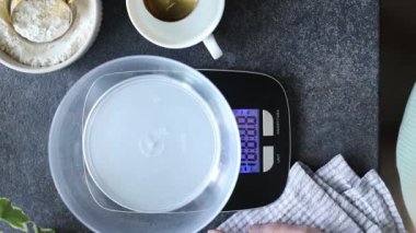 Mutfak terazisini kullanarak kaşıkla su döken bir kadın eli. Evde hamur mayası pişirmek, pişirmek ve beslemek için un tartmak. Teraziler, su ve un mutfak tezgahında. Üst görünüm