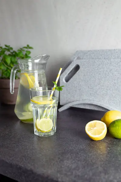 Sürahi ve bardakla ferahlatıcı narenciye ya da mutfak tezgahında kokteyl. Limonata veya mojito hazırlamak için dilimlenmiş limonlar, saksıda naneli otlar, masada taş kesme tahtası, boşluğu kopyalamak için.