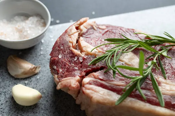 stock image Close-up of fresh raw beef steak garnished with rosemary on cutting board. Salt and garlic cloves on kitchen counter