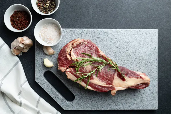 Biberiyeyle süslenmiş taze çiğ biftek. Taş kesme tahtasında. Mutfak tezgahında tuz, karabiber ve sarımsak karanfili.