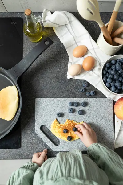 Çocuk eller taş kesme tahtasında krep ve yaban mersiniyle oynuyor. Çeşitli malzemeler - kasede yabanmersini, kesme tahtasında yumurta. Yemek hazırlama, aile zamanı, evde yemek pişirme.