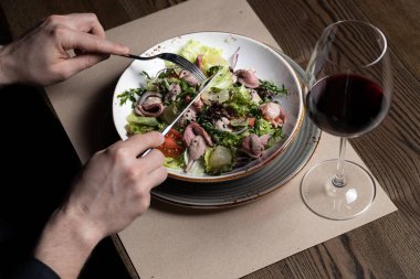 Restoran ortamında kırmızı şarapla salata yiyen biri. Salatada marul, domates ve et var. Salata yemek için çatal ve bıçak kullanan adam.