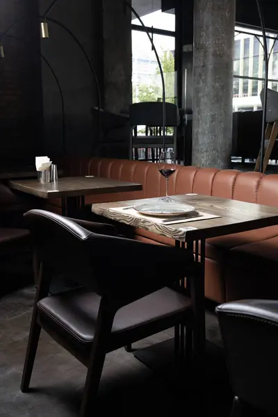 Modern restoranın içinde kahverengi deri koltuklar, ahşap masa, bir bardak şarap, tabak ve gümüş çatal bıçak var. Arka planda geniş pencereli loş ışıklı oda